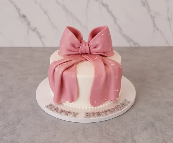 Mujer10 Tierno y elegante pastel redondo con cobertura blanca lisa y un borde inferior de perlas blancas. El gran protagonista del diseño es un enorme moño de fondant rosa con un acabado perlado y efecto de tela drapeada, adornado con diminutas perlas blancas. Sobre la base del pastel se lee el mensaje "HAPPY BIRTHDAY" en letras plateadas. Castillo's Cake Shop.