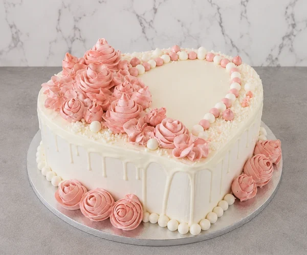 Mujer2 Romántico pastel en forma de corazón con glaseado blanco y un sutil escurrimiento (drip) de chocolate blanco. Está bellamente adornado con una cascada de rosetones de glaseado rosa en distintos tamaños, complementado con un diseño de perlas de azúcar en colores blanco y rosa suave. Castillo's Cake Shop.