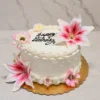 Mujer5 Delicado pastel redondo con glaseado blanco, decorado con bordes clásicos de concha y sutiles perlas blancas. El diseño resalta por la presencia de grandes y realistas flores tipo lirio en tonos blanco y rosa brillante, acompañando el mensaje "Happy Birthday" en el centro con caligrafía negra. Castillo's Cake Shop.