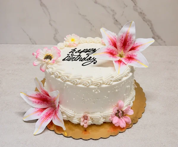 Mujer5 Delicado pastel redondo con glaseado blanco, decorado con bordes clásicos de concha y sutiles perlas blancas. El diseño resalta por la presencia de grandes y realistas flores tipo lirio en tonos blanco y rosa brillante, acompañando el mensaje "Happy Birthday" en el centro con caligrafía negra. Castillo's Cake Shop.