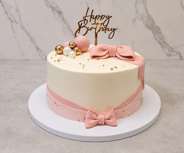 Mujer6 Sofisticado pastel redondo de cobertura blanca, envuelto en su base con una cinta de fondant rosa que termina en un pequeño moño, y coronado con un moño rosa de fondant mucho más grande y estructurado. Está decorado con un elegante arreglo de esferas doradas, blancas y rosas, perlas doradas y un topper de acrílico de "Happy Birthday". Castillo's Cake Shop.