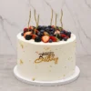 Mujer7 Alto y elegante pastel redondo con glaseado blanco, embellecido con delicados toques de hoja de oro y pequeñas perlas doradas. La parte superior rebosa de una fresca y colorida mezcla de frutos rojos (fresas, zarzamoras y arándanos azules), acompañados de delgadas velas doradas y un letrero frontal dorado de "Happy Birthday". Castillo's Cake Shop.