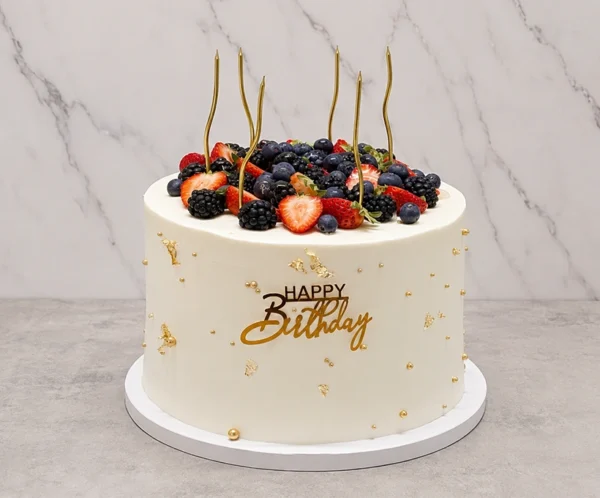 Mujer7 Alto y elegante pastel redondo con glaseado blanco, embellecido con delicados toques de hoja de oro y pequeñas perlas doradas. La parte superior rebosa de una fresca y colorida mezcla de frutos rojos (fresas, zarzamoras y arándanos azules), acompañados de delgadas velas doradas y un letrero frontal dorado de "Happy Birthday". Castillo's Cake Shop.
