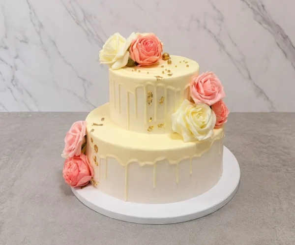 Mujer8 Hermoso pastel de dos pisos con cobertura en tono crema claro y un escurrimiento (drip) de chocolate a juego en ambos niveles. Su decoración es clásica y elegante, conformada por preciosas rosas de glaseado en colores rosa y crema, acentuadas con sutiles destellos de hoja de oro. Castillo's Cake Shop.