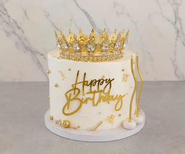 Mujer9 Majestuoso pastel redondo con glaseado blanco, decorado lujosamente con esferas doradas de distintos tamaños, perlas, chispas y toques de hoja de oro. Destaca una impresionante y brillante corona dorada con pedrería en la cima, junto a un letrero acrílico de "Happy Birthday" en el frente y dos velas doradas de diseño ondulado. Castillo's Cake Shop.