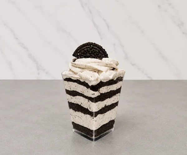 Oreo en vasito. Postre en capas servido en un vasito transparente cuadrado. Intercala gruesas líneas de polvo oscuro de galleta triturada con una crema clara moteada. Se corona con un abundante rosetón de crema sabor cookies and cream y la mitad de una galleta Oreo tradicional.