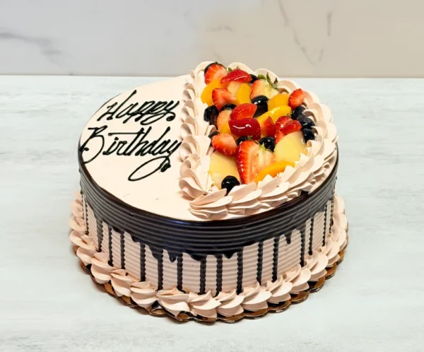 Pastel de Chocolate Tres Leches Pastel redondo con glaseado color moca claro texturizado y un marcado goteo de chocolate oscuro. La superficie está dividida por una línea de rosetones; un lado lleva el mensaje "Happy Birthday" en elegante letra cursiva negra, y el otro está cubierto de frutas frescas mixtas (fresas, duraznos, kiwi y frutos rojos).