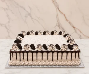 Pastel de formato rectangular con glaseado tipo cookies and cream (con pequeñas motas de galleta triturada), textura estriada y goteo de chocolate oscuro. Todo el perímetro superior está adornado con rosetones de glaseado decorados con finos hilos de chocolate y mitades de galleta Oreo incrustadas.