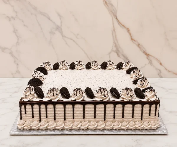 Pastel de Oreo Rectangular Pastel de formato rectangular con glaseado tipo cookies and cream (con pequeñas motas de galleta triturada), textura estriada y goteo de chocolate oscuro. Todo el perímetro superior está adornado con rosetones de glaseado decorados con finos hilos de chocolate y mitades de galleta Oreo incrustadas.