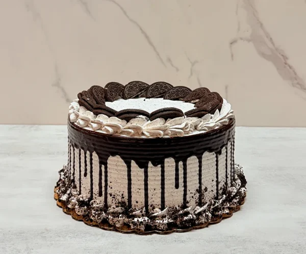 Pastel de Oreo Redondo 0 Pasteles redondos cubiertos con glaseado de cookies and cream con textura de líneas horizontales y un goteo de chocolate oscuro profundo. En la parte superior destacan mitades de galletas Oreo entrelazadas formando un círculo interior, rodeadas por rosetones de glaseado. El borde inferior está rematado con rosetones y migajas de galleta Oreo espolvoreadas.