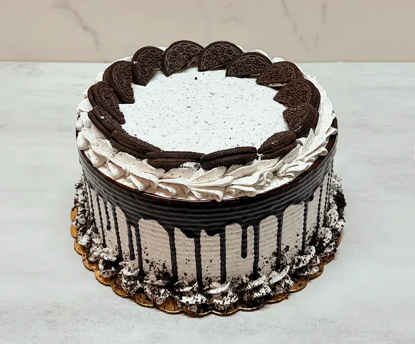 Pastel de Oreo Redondo 1 Pasteles redondos cubiertos con glaseado de cookies and cream con textura de líneas horizontales y un goteo de chocolate oscuro profundo. En la parte superior destacan mitades de galletas Oreo entrelazadas formando un círculo interior, rodeadas por rosetones de glaseado. El borde inferior está rematado con rosetones y migajas de galleta Oreo espolvoreadas.