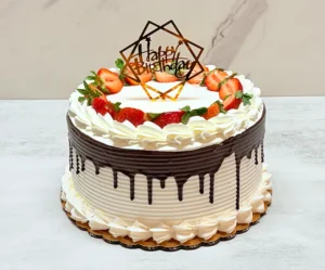 Pastel redondo con glaseado blanco texturizado con finas líneas horizontales y decorado con un atractivo goteo de chocolate oscuro. La parte superior presenta mitades de fresas frescas dispuestas en un anillo perfecto, enmarcadas por rosetones de glaseado blanco, y está coronado en el centro con un elegante adorno geométrico dorado con la frase "Happy Birthday". El borde inferior está cuidadosamente rematado con una hilera continua de rosetones de glaseado blanco.