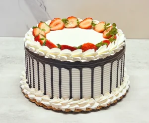 Pastel redondo con glaseado blanco texturizado con finas líneas horizontales y decorado con un atractivo goteo de chocolate oscuro. La parte superior presenta mitades de fresas frescas dispuestas en un anillo perfecto, enmarcadas por rosetones de glaseado blanco, y está coronado en el centro con un elegante adorno geométrico dorado con la frase "Happy Birthday". El borde inferior está cuidadosamente rematado con una hilera continua de rosetones de glaseado blanco.