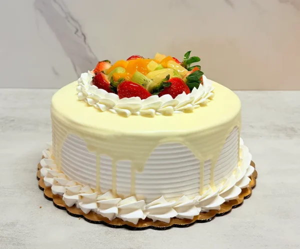 Pastel de Tres Leches 2 Pastel redondo cubierto con glaseado blanco de textura estriada y un delicado goteo continuo de chocolate blanco o ganache. En el centro superior destaca una colorida y abundante mezcla de frutas frescas (fresas, kiwi y durazno), rodeada por un borde clásico de rosetones de glaseado blanco.