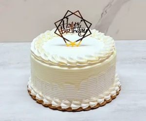 Elegante pastel redondo con glaseado blanco texturizado y un goteo blanco a tono. Su decoración es limpia y minimalista, destacando rosetones de glaseado perfectos en los bordes superior e inferior, y coronado en el centro con un sofisticado adorno (topper) geométrico dorado con la frase "Happy Birthday".