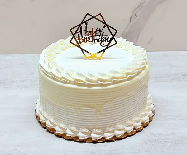 Pastel de Tres Leches sin Fruta Elegante pastel redondo con glaseado blanco texturizado y un goteo blanco a tono. Su decoración es limpia y minimalista, destacando rosetones de glaseado perfectos en los bordes superior e inferior, y coronado en el centro con un sofisticado adorno (topper) geométrico dorado con la frase "Happy Birthday".