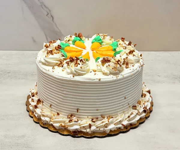Pastel de Zanahoria Pastel redondo con glaseado blanco de líneas horizontales. Su decoración incluye detalles temáticos encantadores: pequeñas zanahorias hechas con glaseado naranja y verde en el centro, complementadas con rosetones blancos y nueces picadas esparcidas tanto en la corona como en el borde inferior.