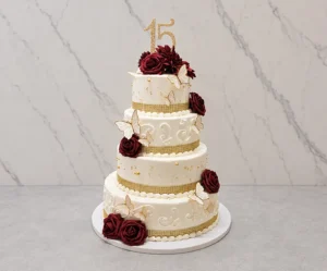 Elegante pastel de XV años de cuatro pisos en color blanco, decorado con sutiles arabescos de glaseado, finas perlas doradas esparcidas y bordes de cinta de pedrería dorada. Embellecido con hermosas rosas color rojo vino, mariposas 3D blancas con siluetas doradas y un topper de "15" dorado brillante en la corona.