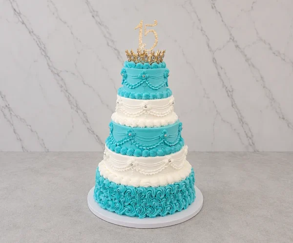 PastelXV_3 Imponente pastel de XV años de cinco pisos que alterna vibrantes colores azul turquesa y blanco. El nivel base presenta rosetones azules, mientras que los demás lucen elegantes drapeados, guirnaldas y bordes de glaseado clásico adornados con perlas plateadas. En la parte superior, destaca una pequeña corona dorada y un adorno de "15" brillante.