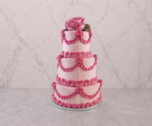 PastelXV_5 Pastel de tres pisos en tono rosa pastel con un exquisito diseño de glaseado estilo Lambeth. Decorado con elaborados drapeados, bordes de concha y detalles entrelazados en un rosa más intenso, acentuados con finos hilos blancos y perlas de azúcar. Rematado en la corona con una hermosa rosa natural.