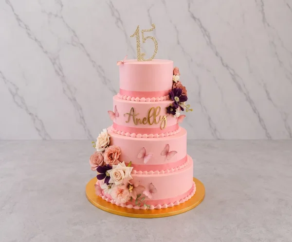 PastelXV_8 Pastel de XV años de cuatro pisos con cobertura lisa en color rosa pastel. Presenta bordes decorados con listón rosa oscuro y perlas de glaseado. Está bellamente adornado con un arreglo floral en cascada en tonos rosa, crema y morado, mariposas 3D, el nombre "Anelly" en letras doradas y un topper de "15" dorado brillante en la parte superior.
