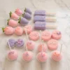 Un hermoso surtido de postres para mesa dulce en delicados tonos pastel rosa y lila. El conjunto incluye fresas cubiertas de chocolate, galletas cubiertas (tipo Oreo) y paletas tipo Magnum (cakesicles), todas decoradas con líneas finas, chispas de colores y aplicaciones de mariposas. En la parte frontal destacan varias cake pops rosas adornadas con perlitas blancas y pequeñas flores de fondant