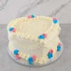 Pastel en forma de corazón con glaseado blanco y decoración estilo Lambeth. Presenta elegantes bordes y guirnaldas acanaladas, adornado con pequeños rosetones en color rosa y azul, ideal para una revelación de género. En el centro superior lleva la palabra "Baby" escrita con glaseado. Castillo's Cake Shop.
