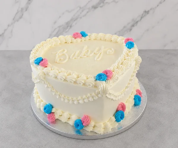 Pastel en forma de corazón con glaseado blanco y decoración estilo Lambeth. Presenta elegantes bordes y guirnaldas acanaladas, adornado con pequeños rosetones en color rosa y azul, ideal para una revelación de género. En el centro superior lleva la palabra "Baby" escrita con glaseado. Castillo's Cake Shop.