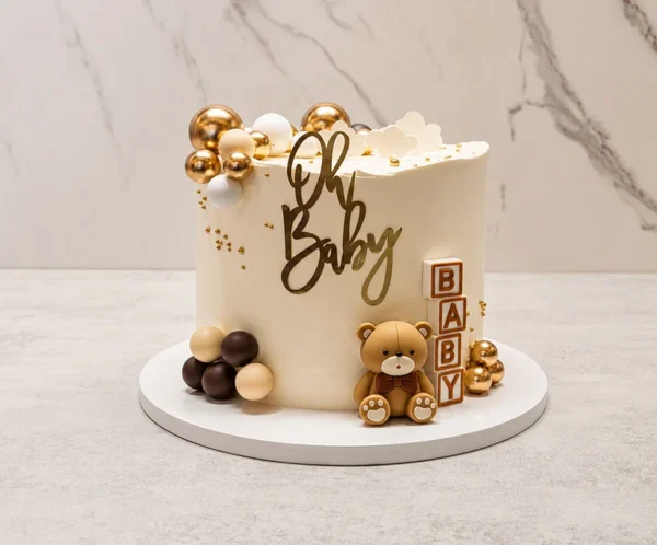 Pastel redondo de un piso con cobertura lisa en tono crema. Está decorado con un tierno osito de fondant, cubos de juguete que forman la palabra "BABY", y un hermoso arreglo de esferas de chocolate en tonos dorado metálico, blanco y café. En la parte superior luce un letrero dorado de "Oh Baby". Castillo's Cake Shop.