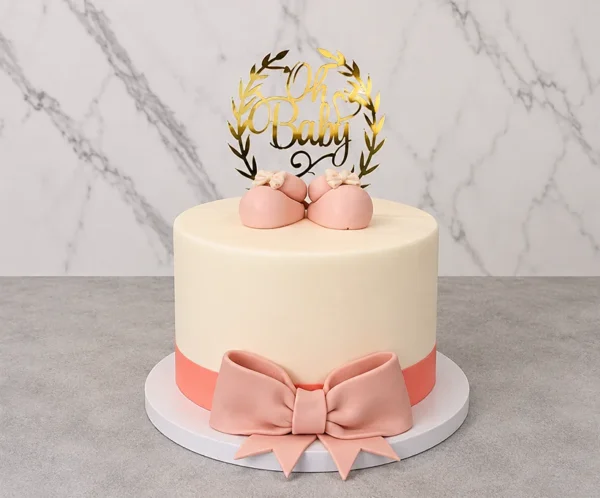 Elegante pastel redondo de un piso con cobertura lisa en color crema. En la base destaca un listón y un gran moño de fondant en color rosa viejo. Está coronado con un par de tiernos zapatitos de bebé en fondant rosa y un delicado adorno dorado con corona de laureles que dice "Oh Baby". Castillo's Cake Shop.
