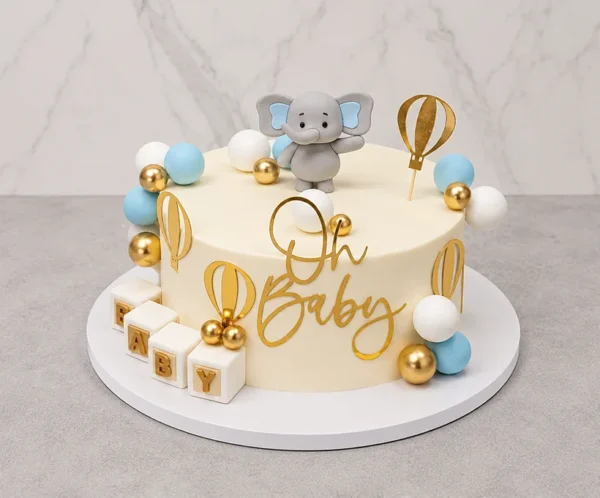 Pastel redondo de un piso con base crema, decorado con una tierna temática de elefantito. Incluye un arreglo de esferas en tonos azul claro, blanco y dorado, siluetas doradas de globos aerostáticos, cubos con las letras "B A B Y", un elefantito de fondant en la parte superior y un letrero dorado de "Oh Baby". Castillo's Cake Shop.