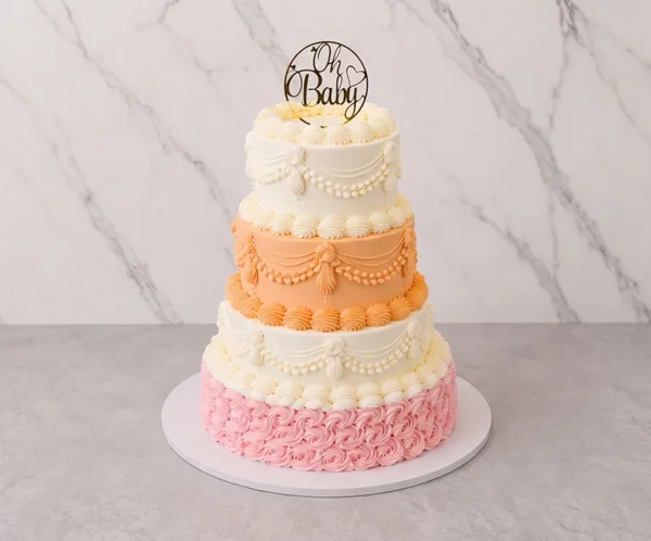Majestuoso pastel de cuatro pisos con decoración de glaseado estilo Lambeth. El piso inferior está completamente cubierto de rosetones rosas, el segundo es de color melocotón con hermosas guirnaldas, y los dos superiores son blancos con intrincados bordes y drapeados clásicos. Rematado con un letrero dorado circular de "Oh Baby". Castillo's Cake Shop.