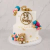 Tierno pastel de dos pisos con cobertura blanca lisa, adornado con sutiles destellos de hoja de oro y racimos de esferas en colores turquesa, rosa, magenta y dorado. Incluye dos ositos de fondant, uno durmiendo plácidamente en la cima y otro sentado en la base sosteniendo un corazón rosa. Destaca un adorno dorado de "Oh Baby" en el frente del piso superior. Castillo's Cake Shop.