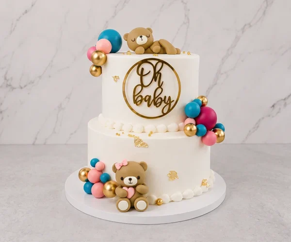 Tierno pastel de dos pisos con cobertura blanca lisa, adornado con sutiles destellos de hoja de oro y racimos de esferas en colores turquesa, rosa, magenta y dorado. Incluye dos ositos de fondant, uno durmiendo plácidamente en la cima y otro sentado en la base sosteniendo un corazón rosa. Destaca un adorno dorado de "Oh Baby" en el frente del piso superior. Castillo's Cake Shop.