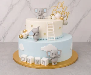 Pastel de dos pisos con temática de elefantitos. El piso inferior es color azul cielo pastel con nubes blancas y cubos que dicen "B A B Y". El piso superior es blanco con estrellas doradas y una escalerita blanca apoyada en el borde. Está decorado con tres adorables elefantes grises de fondant en diferentes posiciones, esferas en tonos blanco, gris y dorado, y un letrero superior de "Oh Baby". Castillo's Cake Shop.